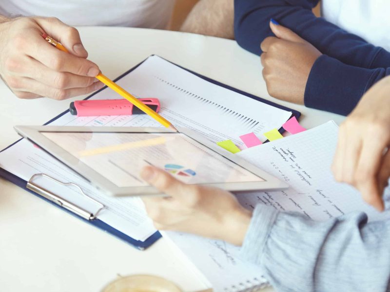 Working in team: young businesspeople discussing business plans and ideas, analyzing graphics and diagrams on paper. Man holding pen, pointing at tablet pc screen, explaining project strategy